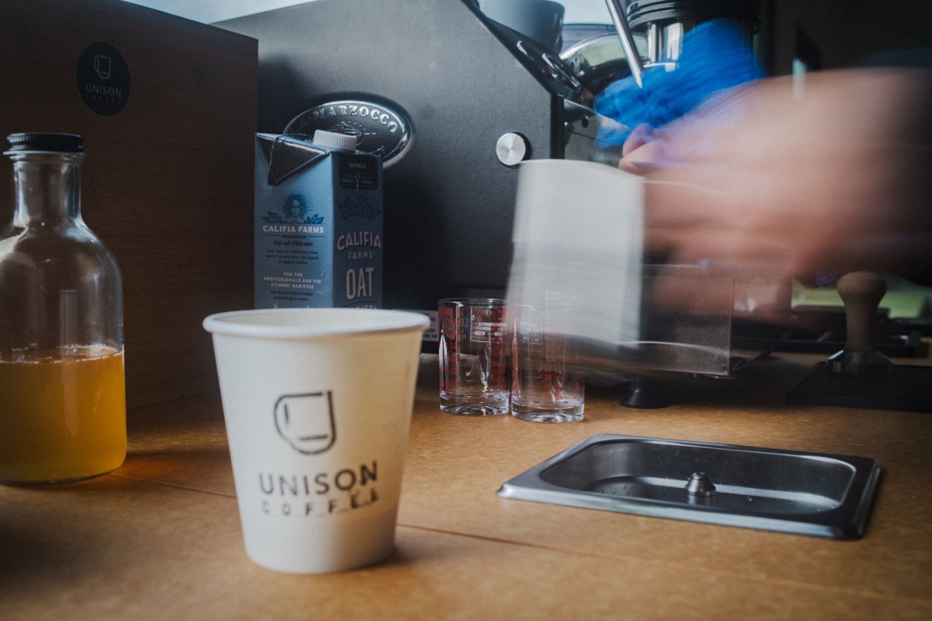 Coffee station with Unison Coffee cup, oat milk, and espresso machine in the background.
